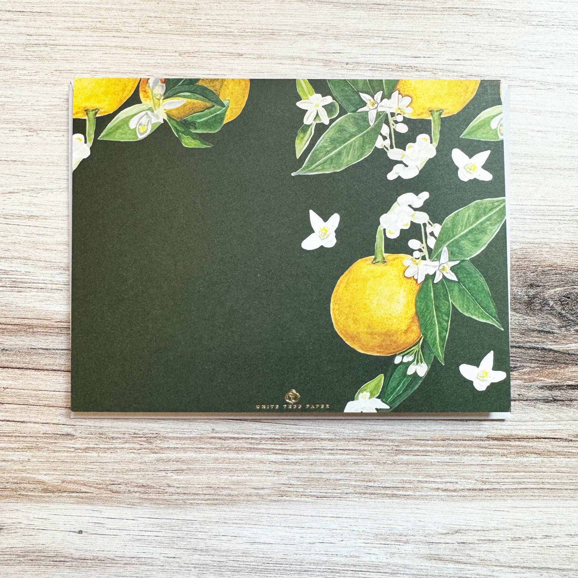 Set of 6 folded full color gold foil stamped cards with dark green background and watercolor painted orange blossom flowers and oranges in foreground, blank inside. 6 luxe gray envelopes. Pictured here is back of card design and White Tree Paper logo.